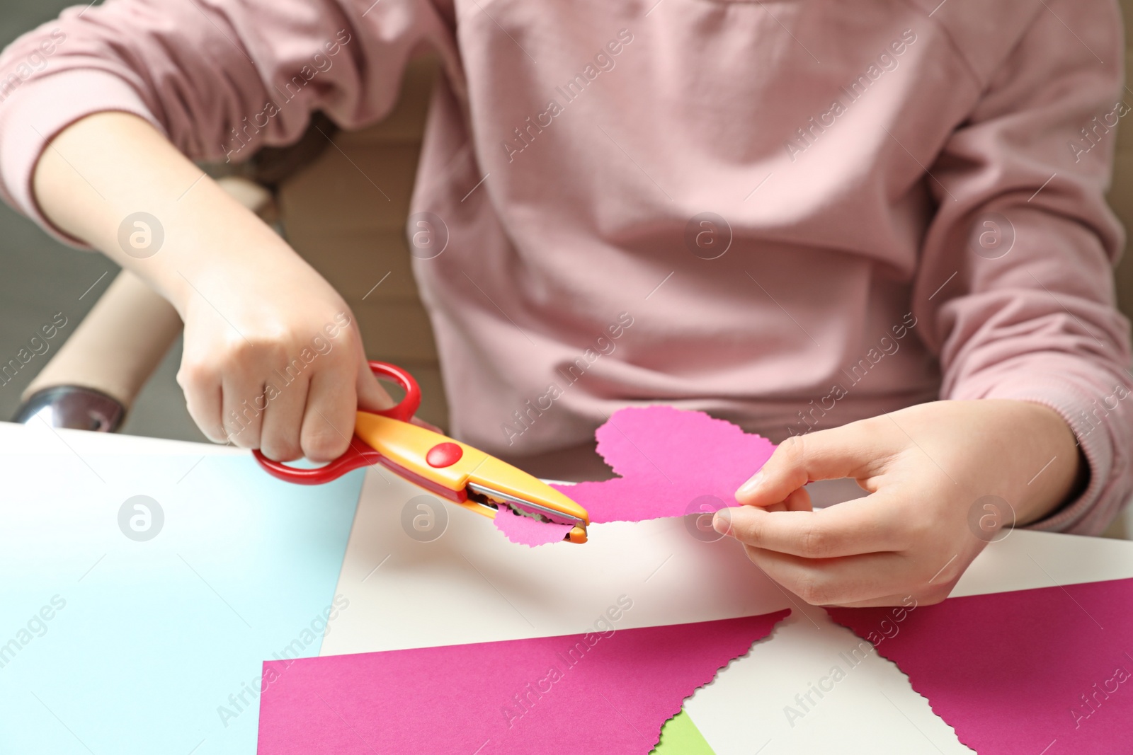 Photo of Child cutting out paper heart with craft scissors at table, closeup