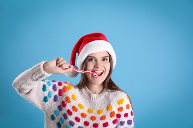 Photo of Pretty woman in Santa hat and festive sweater eating candy cane on light blue background