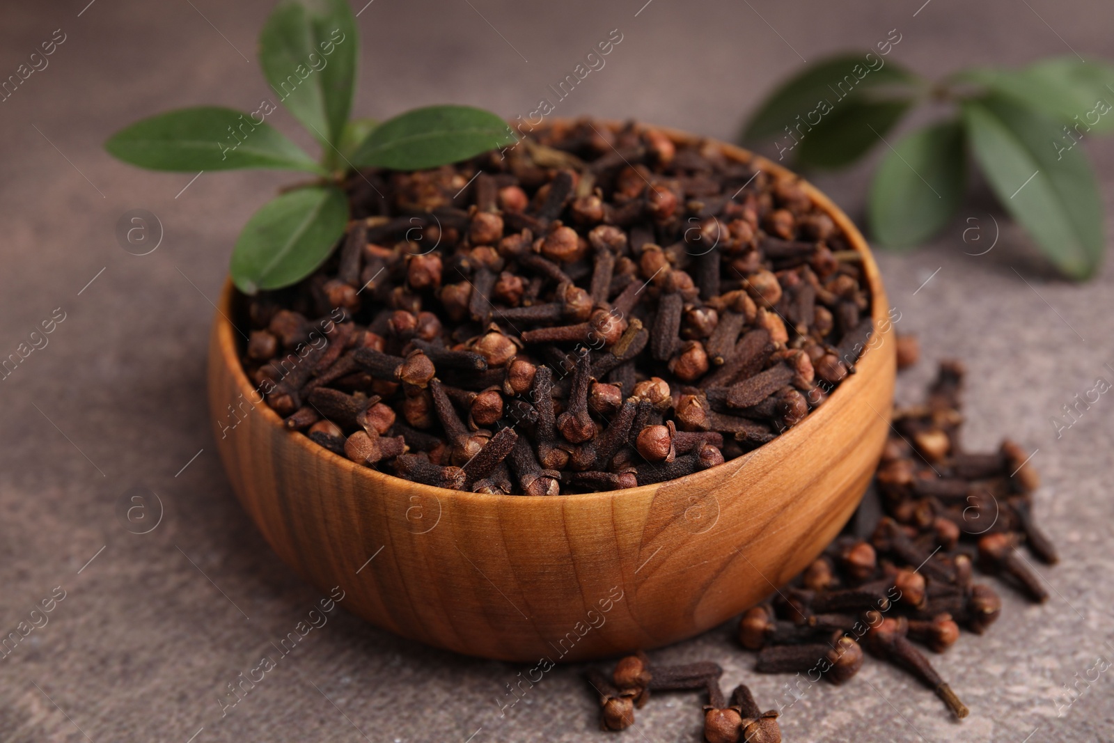 Photo of Aromatic cloves and green leaves in bowl on brown table, closeup