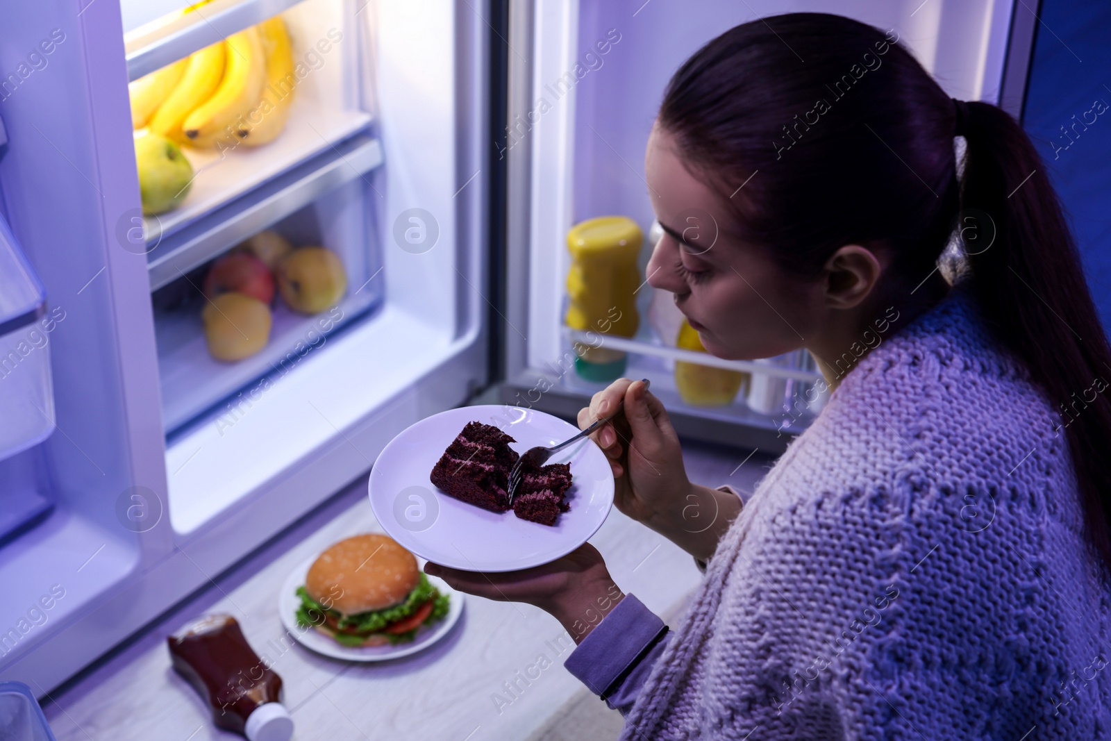 Photo of Young woman eating cake near refrigerator in kitchen at night. Bad habit
