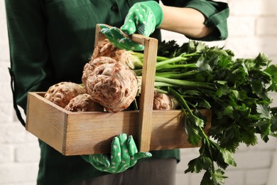 Photo of Woman holding wooden crate with raw celery roots near white brick wall, closeup