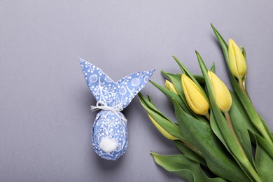 Photo of Easter bunny made of wrapping paper and egg near beautiful tulips on grey background, flat lay. Space for text