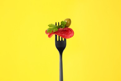 Fork with tasty slice of salami, olive and parsley on yellow background