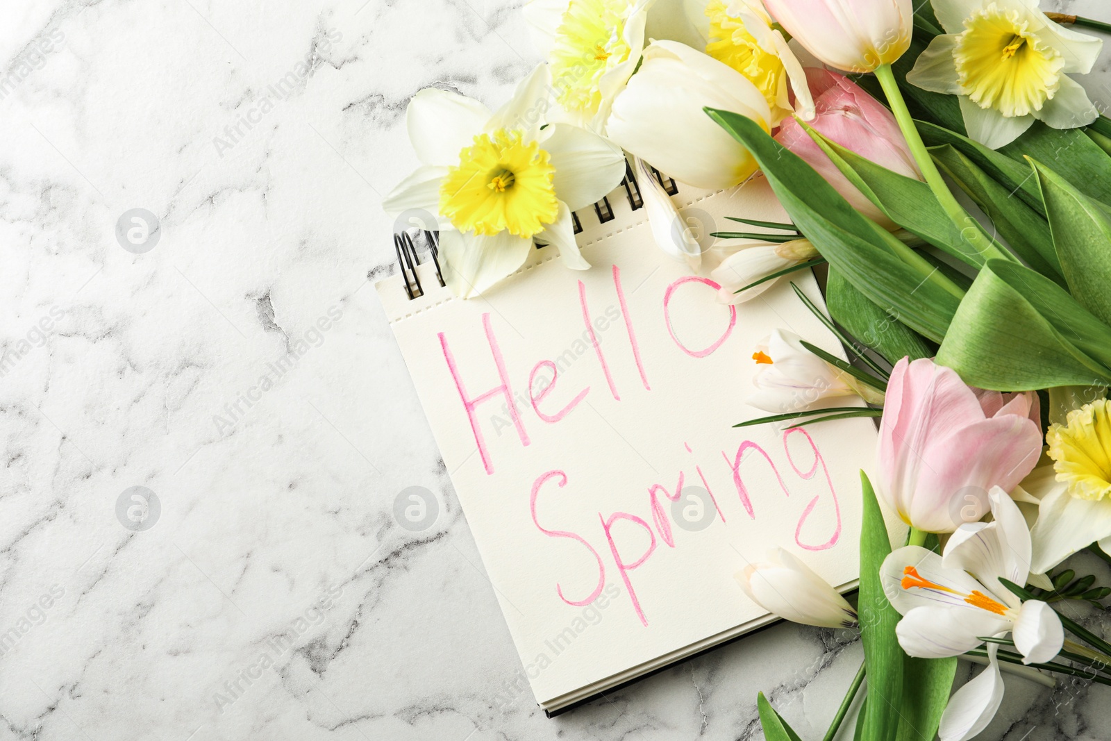 Photo of Notebook with words HELLO SPRING and fresh flowers on white marble table, flat lay