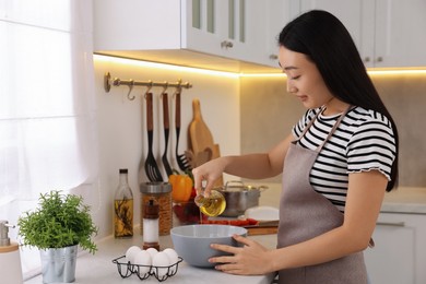 Photo of Cooking process. Beautiful woman pouring oil from bottle into bowl in kitchen