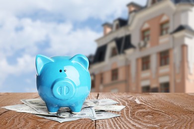 Image of Piggy bank and money on wooden surface and blurred view of beautiful house, space for text. Mortgage concept