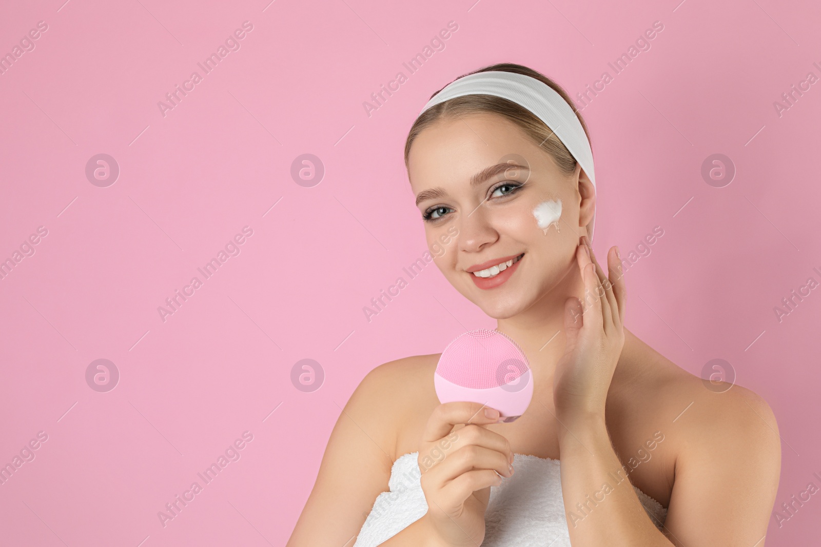 Photo of Young woman washing face with brush and cleansing foam on pink background, space for text. Cosmetic product