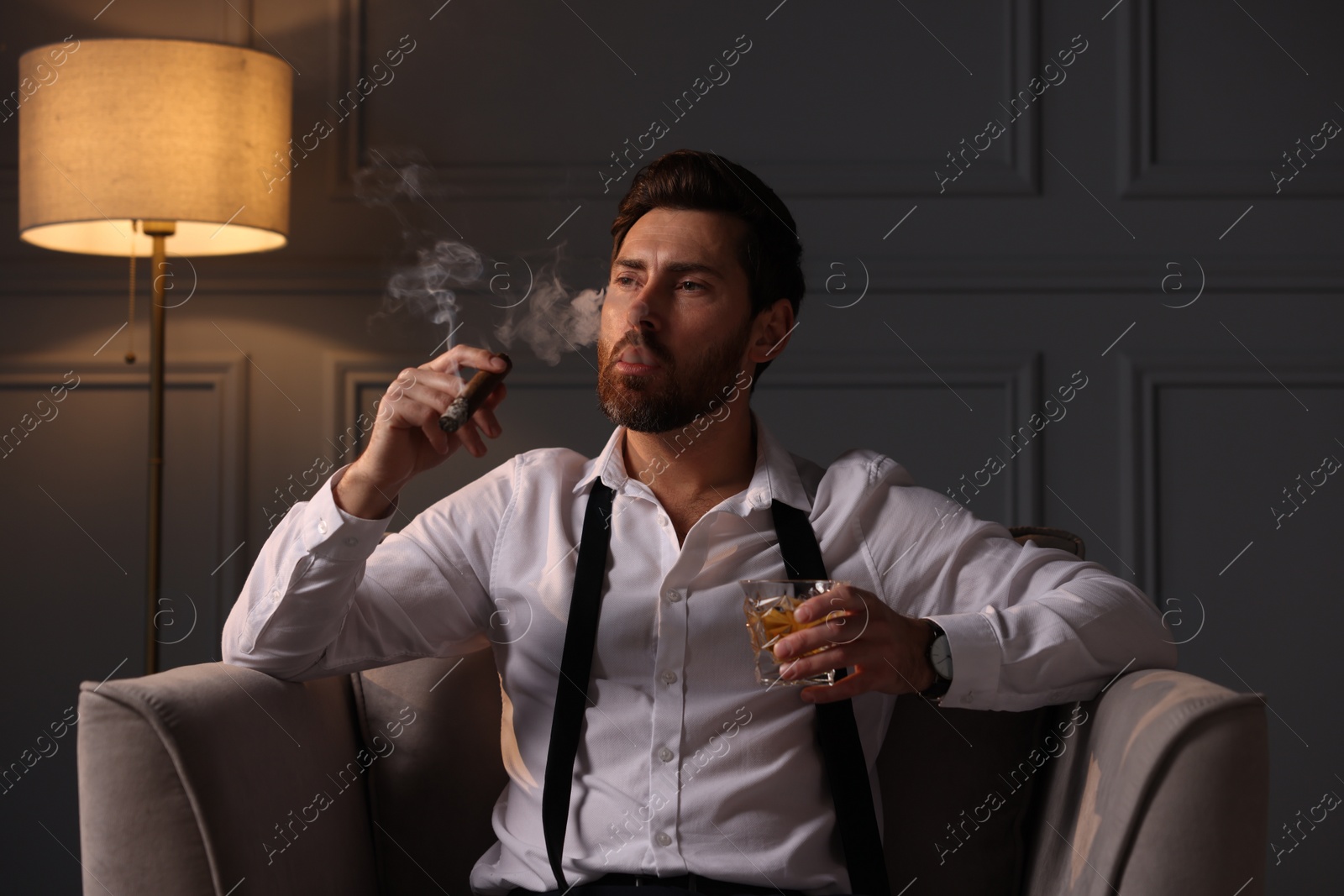 Photo of Handsome man with glass of whiskey smoking cigar in armchair at home