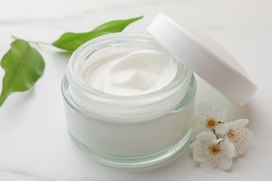 Glass jar of face cream, leaves and flowers on white marble table, closeup