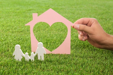 Photo of Man holding cut paper house near figure of family on green grass, closeup