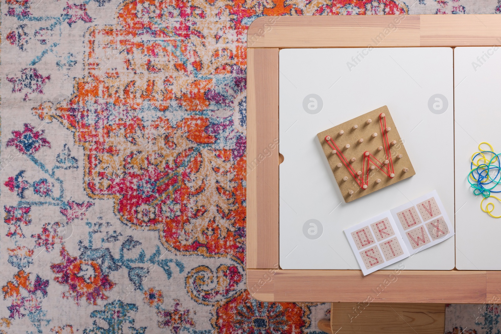 Photo of Wooden geoboard with letter W made of rubber bands and activity book on white table in room, top view with space for text. Motor skills development