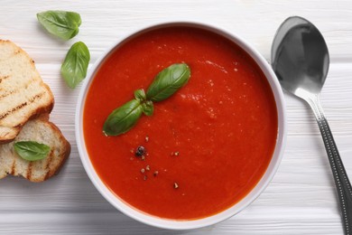 Delicious tomato cream soup in bowl served with pieces of grilled bread on white wooden table, flat lay