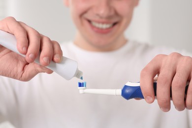 Man squeezing toothpaste from tube onto electric toothbrush on blurred background, closeup