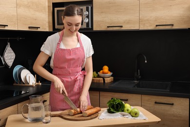Photo of Young woman in striped apron cutting baguette on wooden table at kitchen