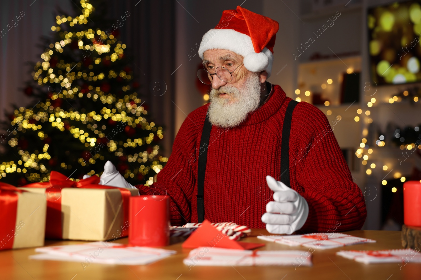 Photo of Santa Claus at his workplace. Letters and gift boxes on table in room with Christmas tree