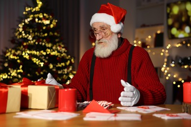 Photo of Santa Claus at his workplace. Letters and gift boxes on table in room with Christmas tree