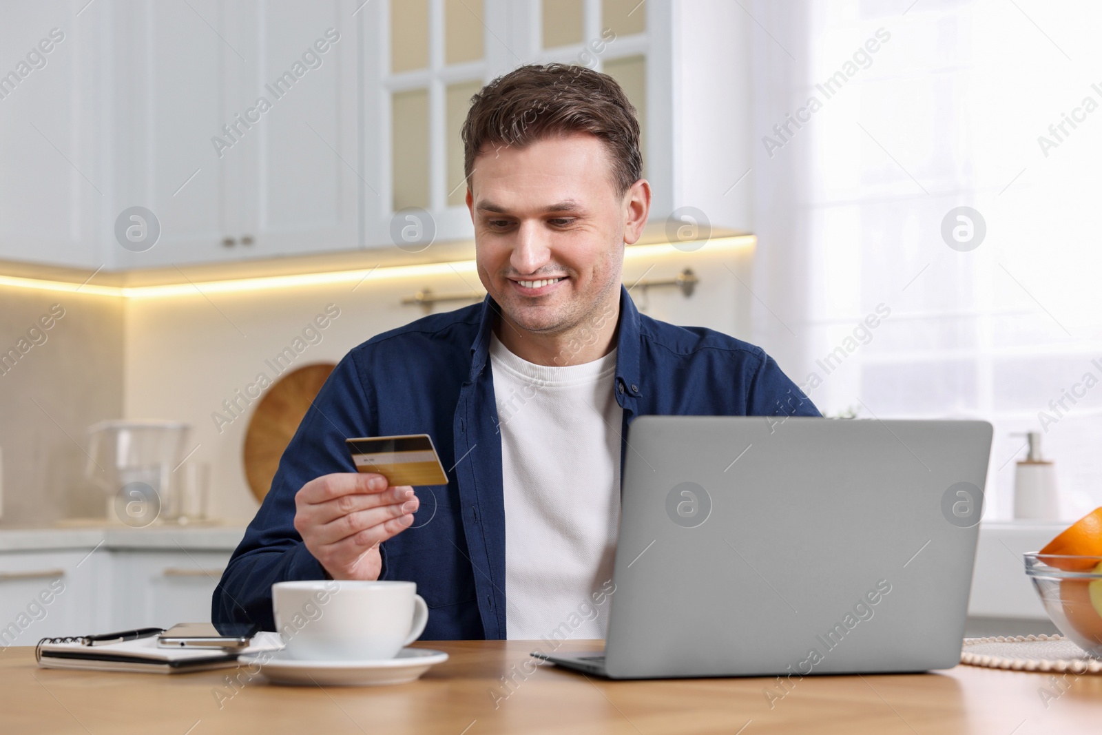 Photo of Happy man with credit card and laptop shopping online at wooden table in kitchen