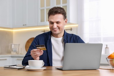 Photo of Happy man with credit card and laptop shopping online at wooden table in kitchen