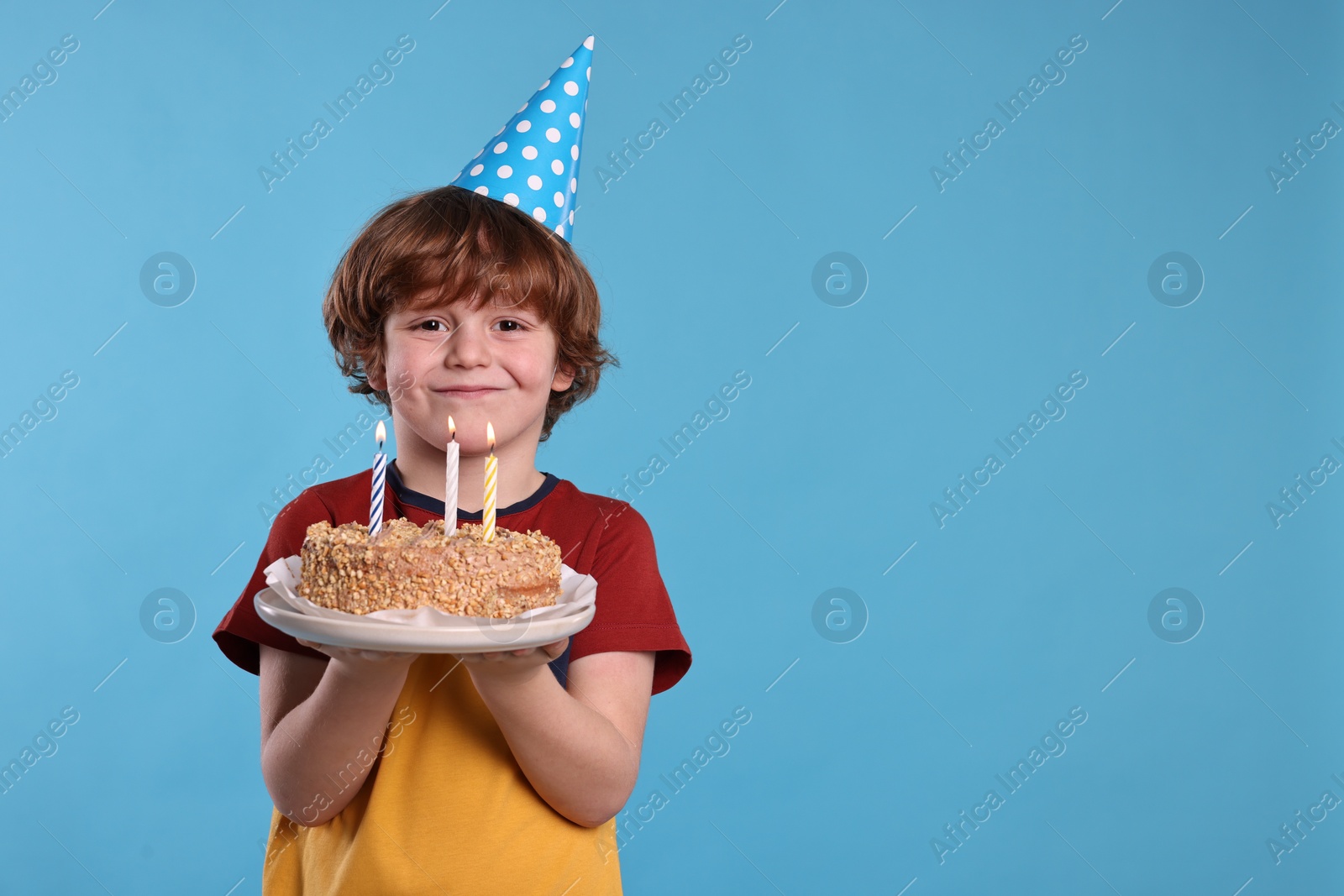 Photo of Birthday celebration. Cute little boy in party hat holding tasty cake with burning candles on light blue background, space for text