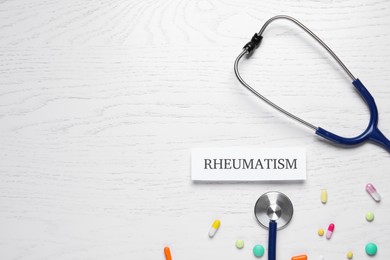 Photo of Card with word Rheumatism, stethoscope and pills on white wooden table, flat lay. Space for text