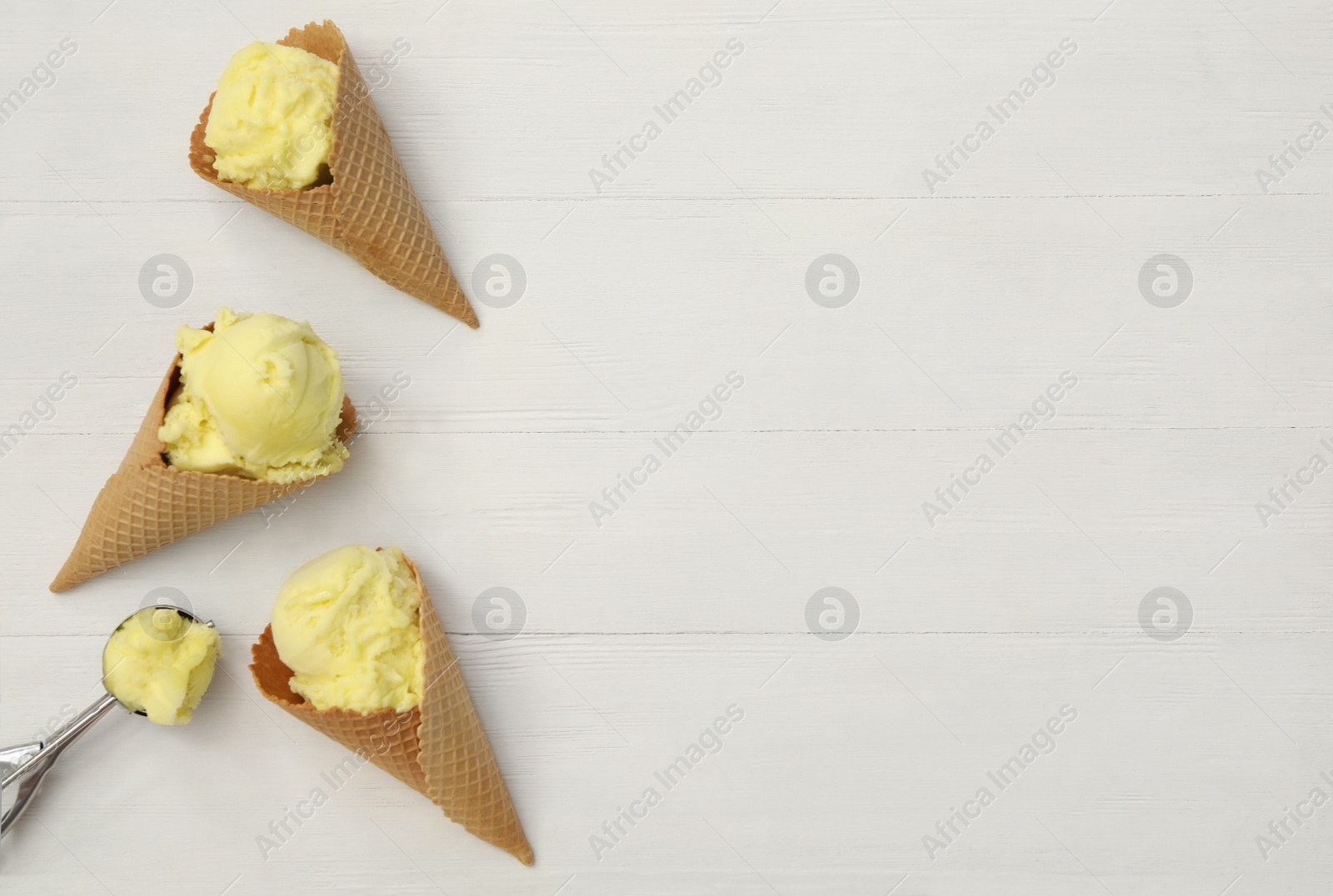 Photo of Yellow ice cream in wafer cones and scoop on white wooden table, flat lay. Space for text