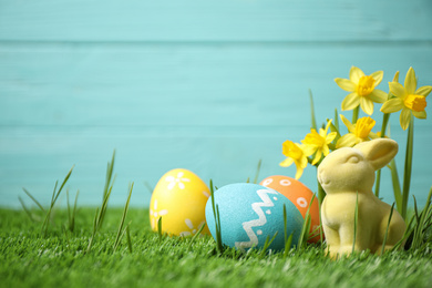 Photo of Colorful Easter eggs, rabbit and narcissus flowers in green grass against light blue background. Space for text