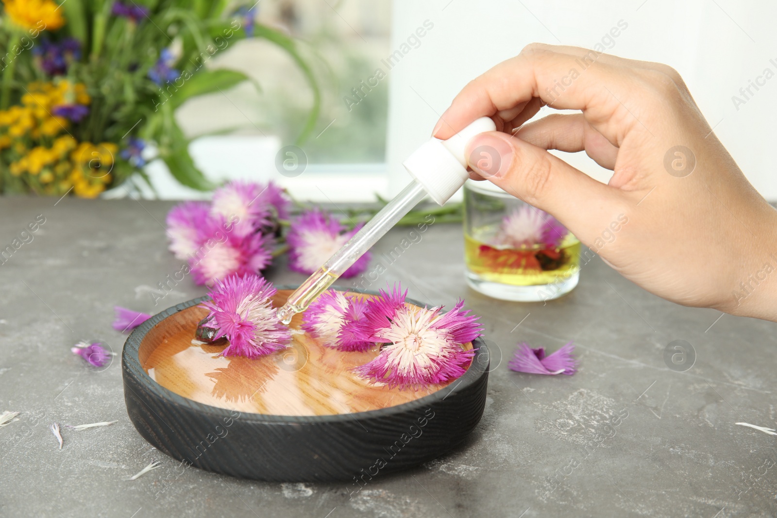 Photo of Woman dripping essential oil into bowl with water and flowers on table
