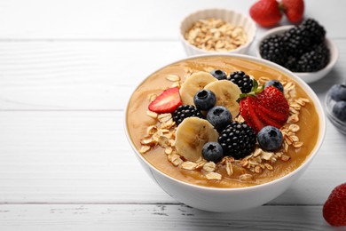 Photo of Delicious smoothie bowl with fresh berries, banana and oatmeal on white wooden table. Space for text