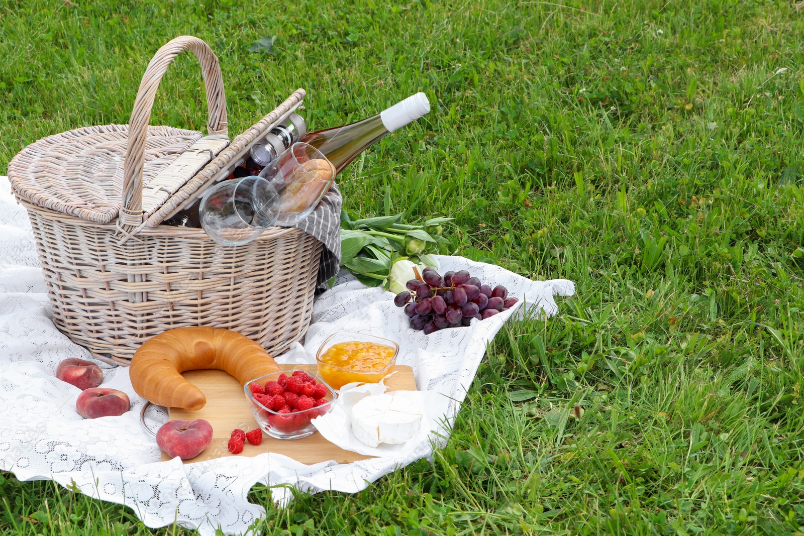 Photo of Picnic basket with tasty food, flowers and cider on blanket outdoors