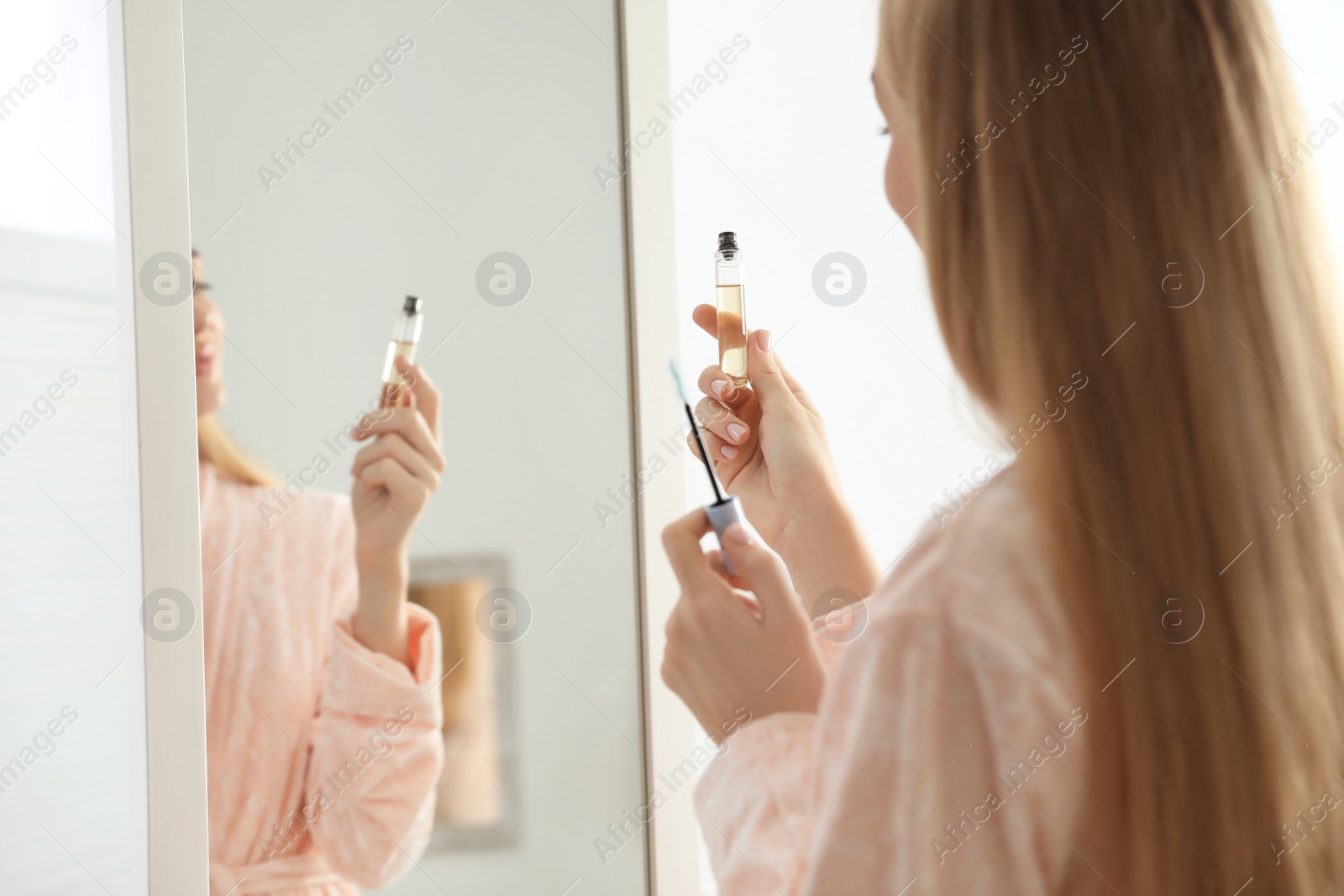 Photo of Young woman applying oil onto her eyelashes near mirror indoors