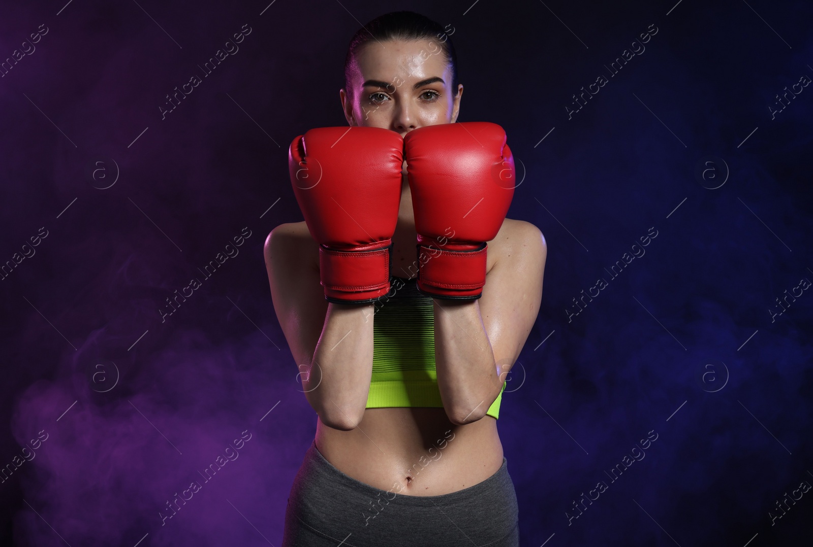 Photo of Portrait of beautiful woman wearing boxing gloves in color lights and smoke on black background