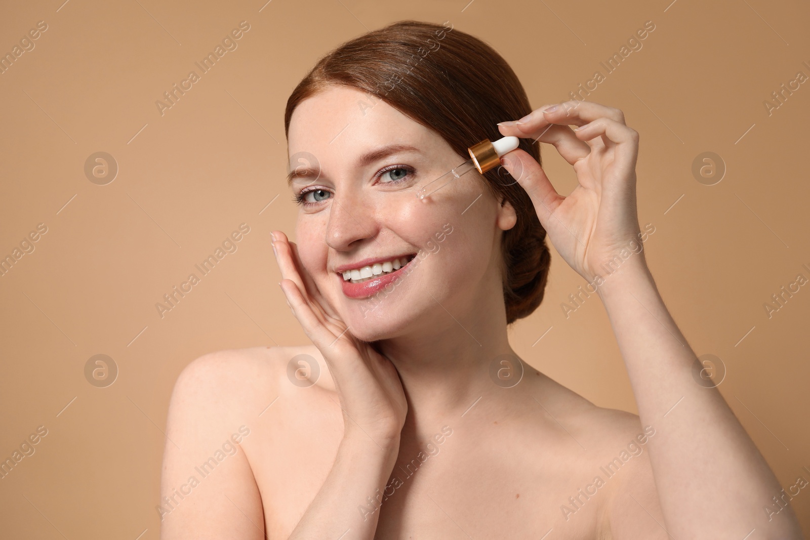 Photo of Smiling woman with freckles applying cosmetic serum onto her face on beige background