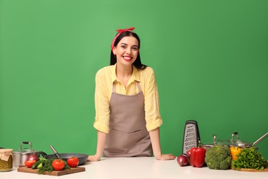 Photo of Young housewife at white table with vegetables and different utensils on green background