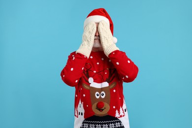 Senior man in Christmas sweater and Santa hat covering face with hands in knitted mittens on light blue background