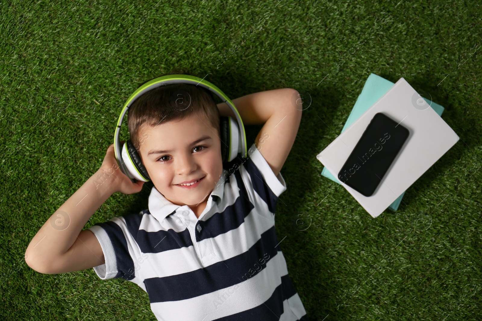 Photo of Cute little boy listening to audiobook on grass, top view