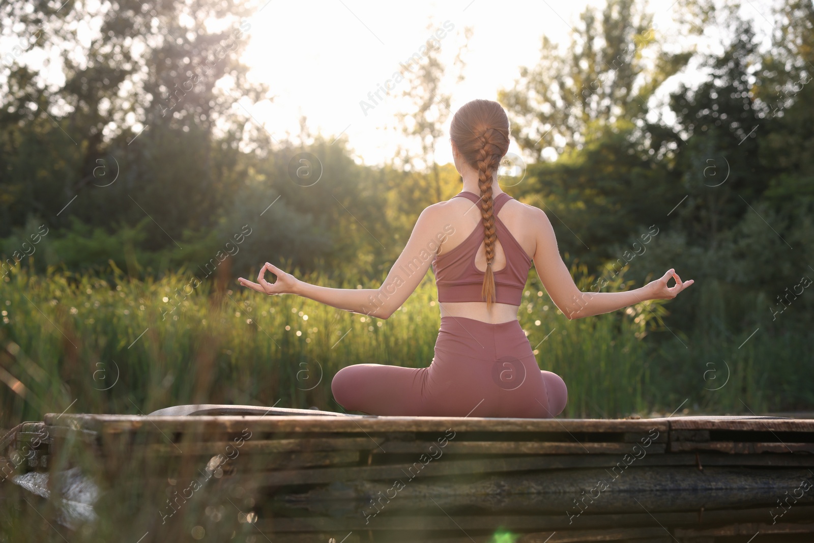 Photo of Woman practicing Padmasana on yoga mat on wooden pier near pond, back view. Lotus pose