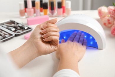 Photo of Woman using ultraviolet lamp to dry gel nail polish at white table, closeup