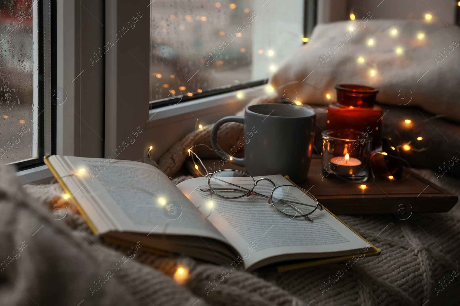 Photo of Cup of hot drink, open book and Christmas lights on knitted blanket near window. Cozy season