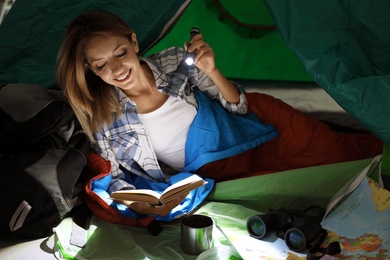 Young woman in sleeping bag reading book with flashlight inside of tent