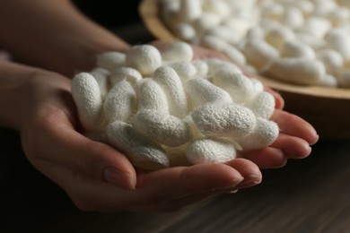 Photo of Woman holding white silk cocoons over bowl at wooden table, closeup