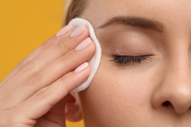 Beautiful woman removing makeup with cotton pad on orange background, closeup