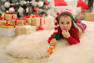 Photo of Cute little girl eating delicious tangerine in room decorated for Christmas