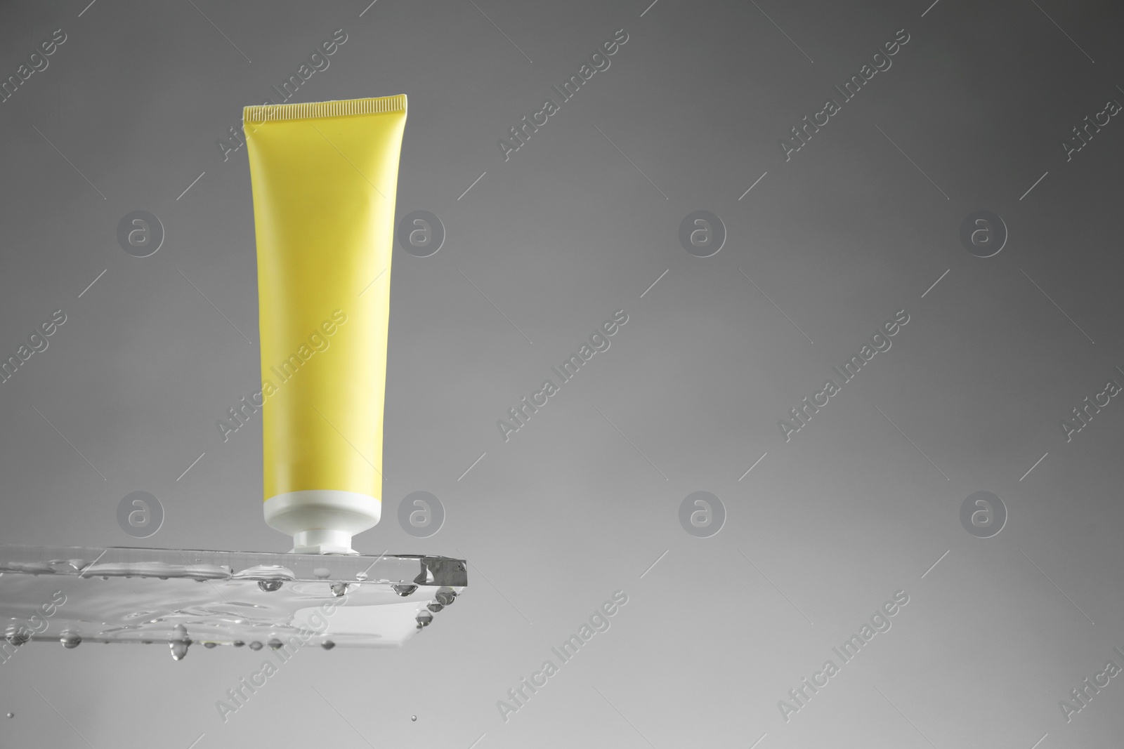 Photo of Moisturizing cream in tube on glass with water drops against grey background, low angle view. Space for text