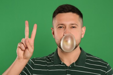 Photo of Handsome man blowing bubble gum and showing peace gesture on green background
