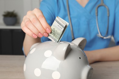 Photo of Doctor putting banknote into piggy bank at wooden table in hospital, closeup. Medical insurance