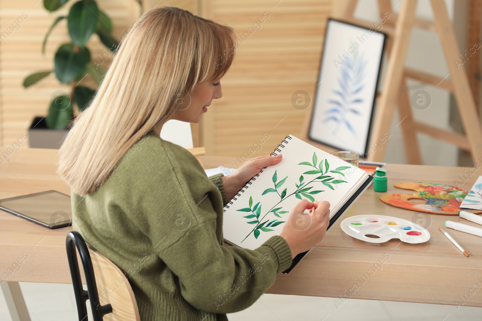 Photo of Young woman painting green twig in sketchbook at wooden table indoors