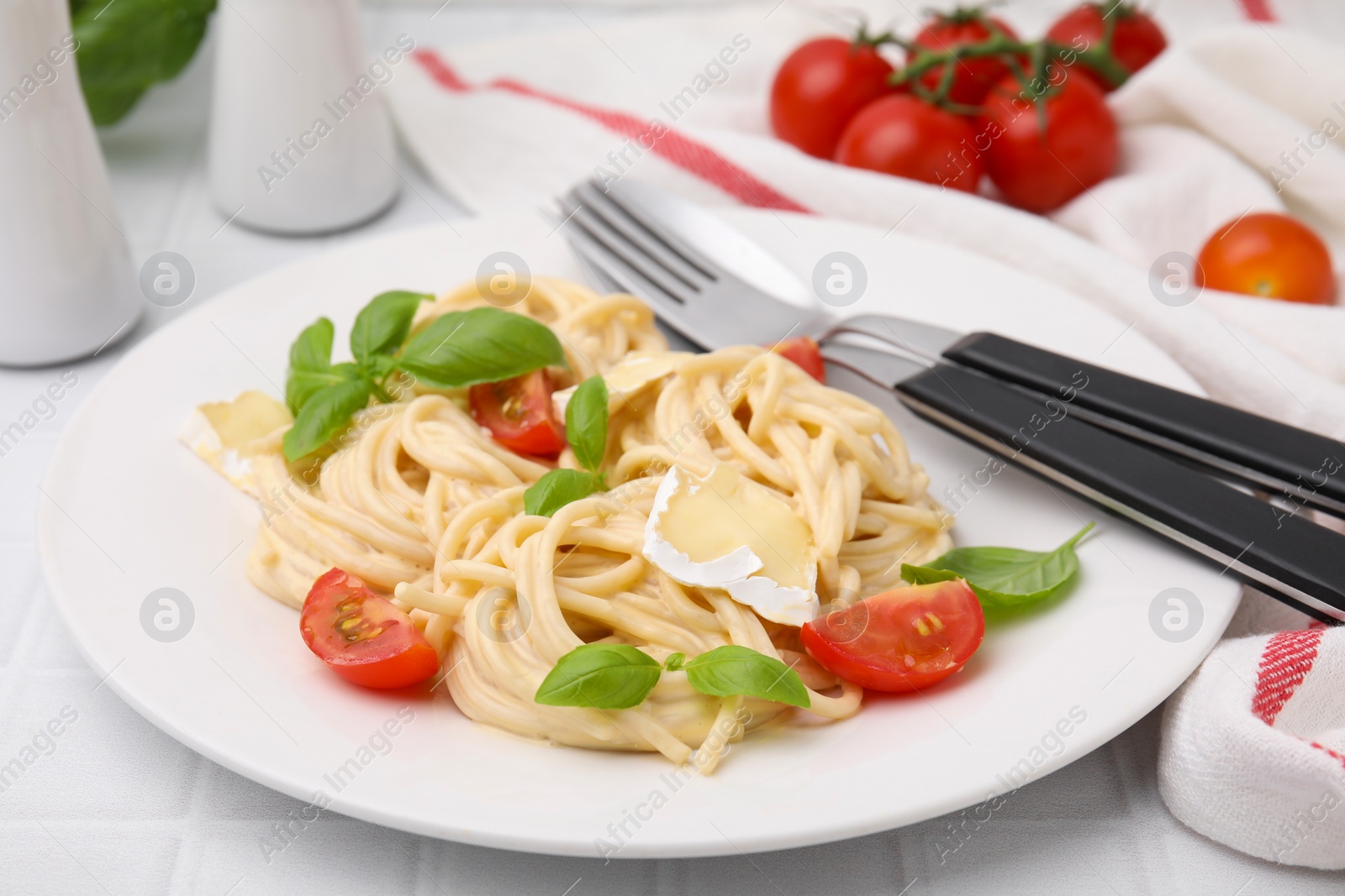 Photo of Delicious pasta with brie cheese, tomatoes, basil and cutlery on white tiled table, closeup