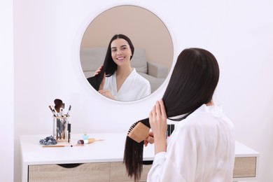 Photo of Beautiful young woman brushing hair while looking in mirror at dressing table indoors