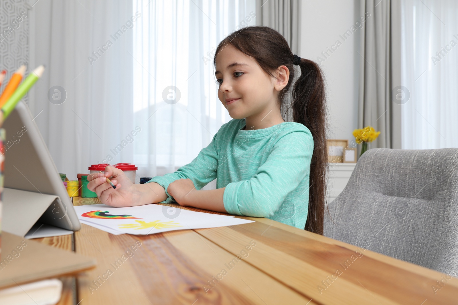 Photo of Little girl drawing on paper with paints at online lesson indoors. Distance learning