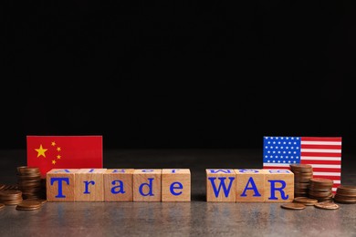 Photo of Words Trade War made of wooden cubes, American and Chinese flags with coins on grey table